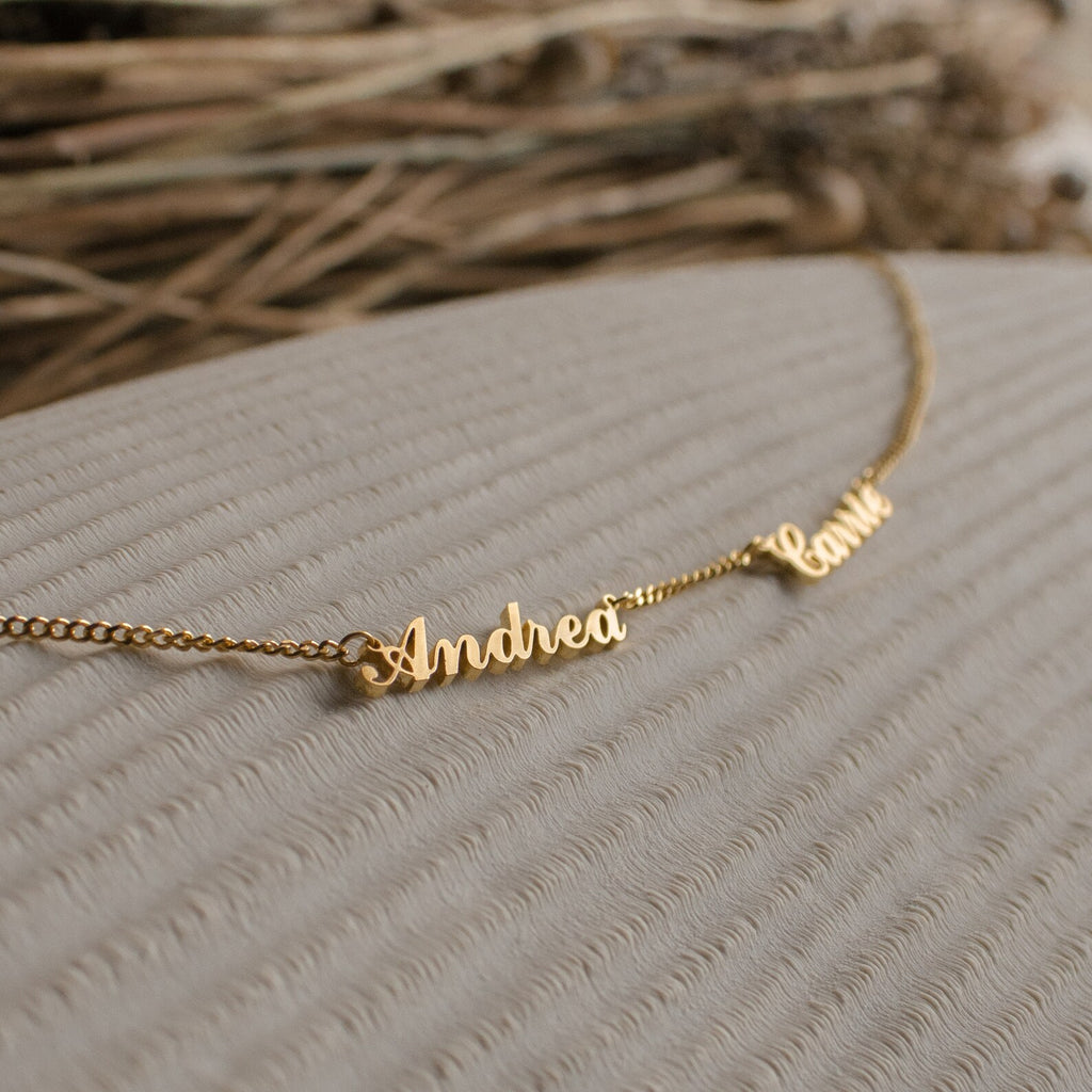 Carrie Multiple Name Necklace in Curb Chain with cursive name charms Andrea and Camila displayed on a textured beige surface—a stunning example of personalized jewelry.