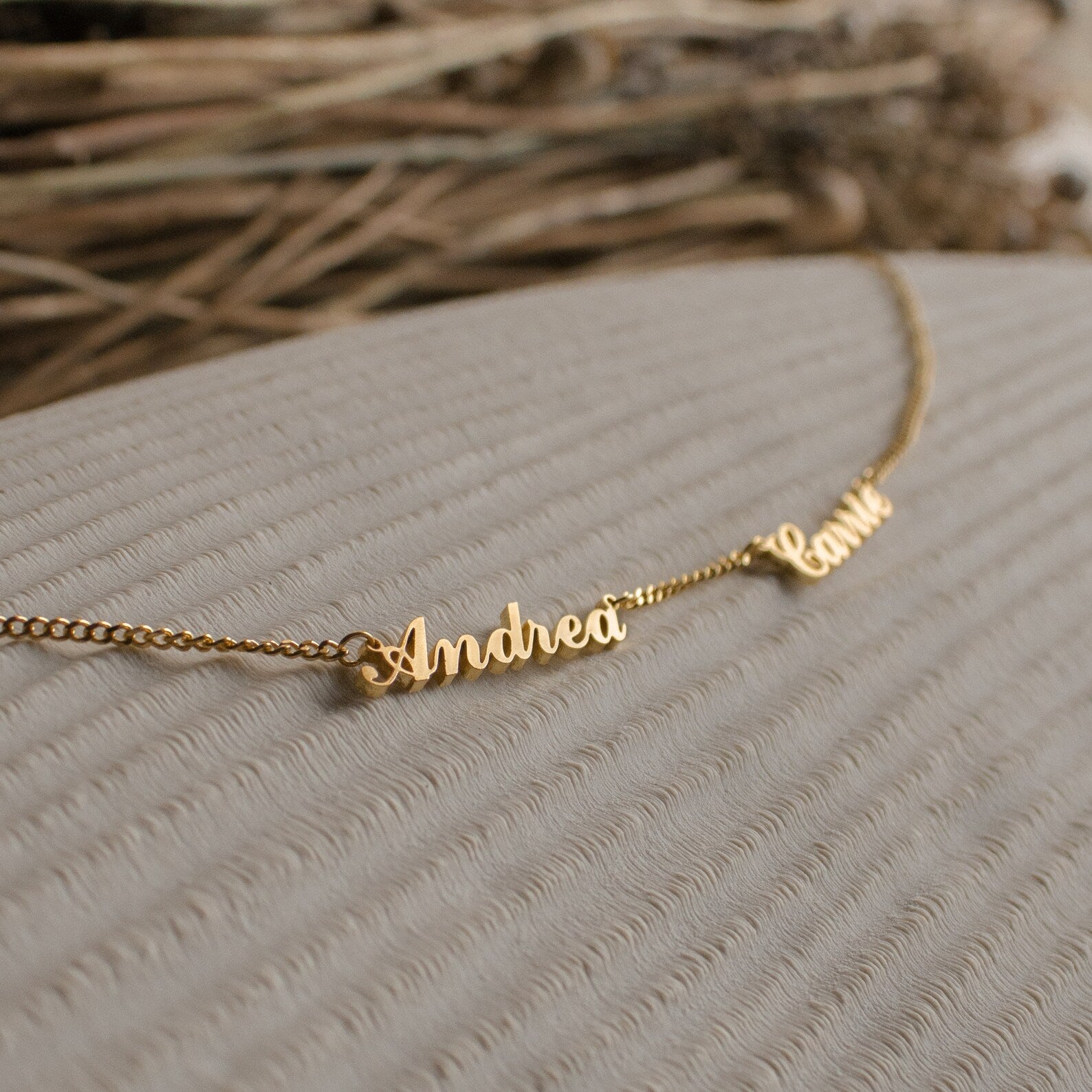 Carrie Multiple Name Necklace in Curb Chain with cursive name charms Andrea and Camila displayed on a textured beige surface—a stunning example of personalized jewelry.