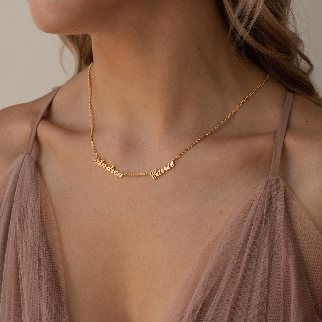 A woman wears a Carrie Multiple Name Necklace in Curb Chain and a sheer, blush-colored dress with thin straps, highlighting her love for personalized jewelry.