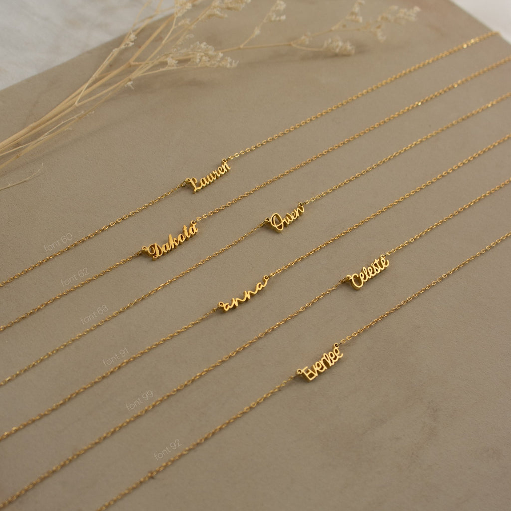 Six Tiny London Name Necklaces are displayed on a beige surface with dried flowers behind them, each featuring a customizable name plate for a unique and minimalist look.