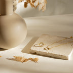 The Kayla Pave Circle Necklace is shown resting on a stone tile beside a gold ring, a beige vase, and dried leaves in gentle sunlight.