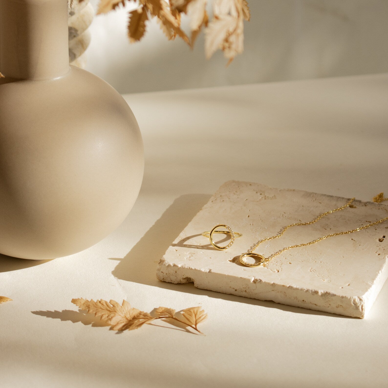 The Kayla Pave Circle Necklace is shown resting on a stone tile beside a gold ring, a beige vase, and dried leaves in gentle sunlight.