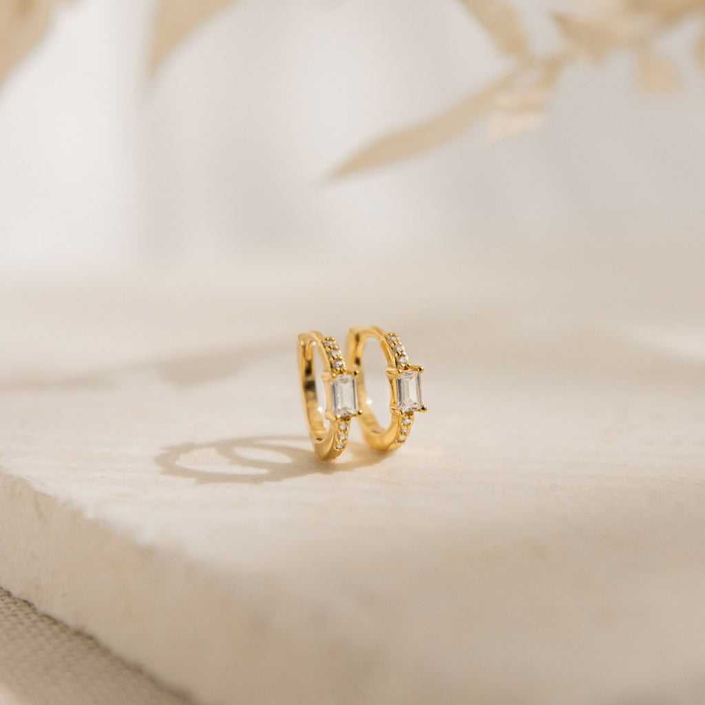 The Natalie Pave Huggies are dainty hoop earrings with rectangular clear gemstones, reminiscent of baguette diamond earrings, displayed on a light textured surface.