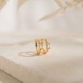 The Natalie Pave Huggies are dainty hoop earrings with rectangular clear gemstones, reminiscent of baguette diamond earrings, displayed on a light textured surface.