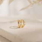 The Natalie Pave Huggies are dainty hoop earrings with rectangular clear gemstones, reminiscent of baguette diamond earrings, displayed on a light textured surface.