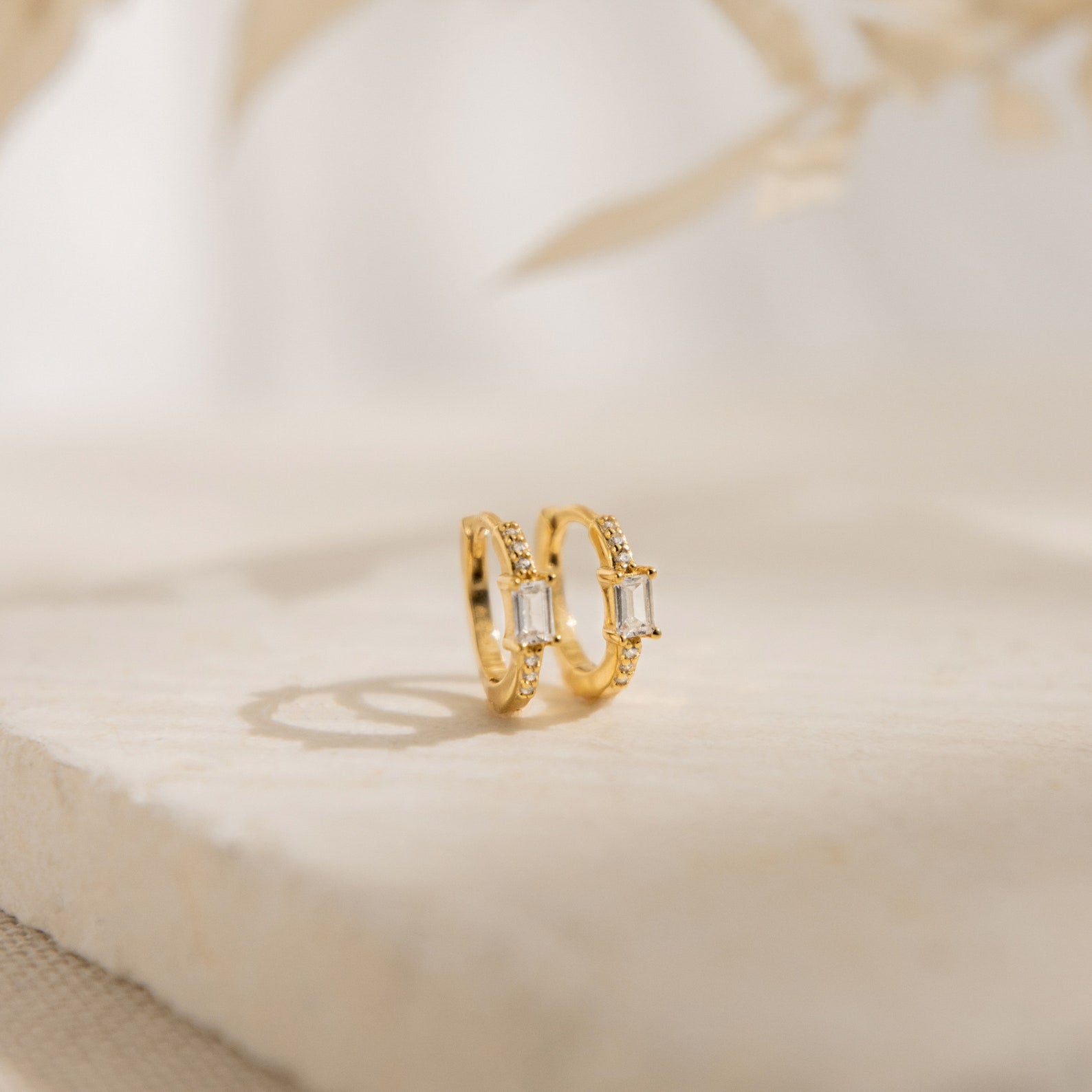 The Natalie Pave Huggies are dainty hoop earrings with rectangular clear gemstones, reminiscent of baguette diamond earrings, displayed on a light textured surface.