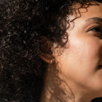 Close-up of a woman with curly hair wearing Natalie Pave Huggies, sunlight casting gentle shadows on her face.