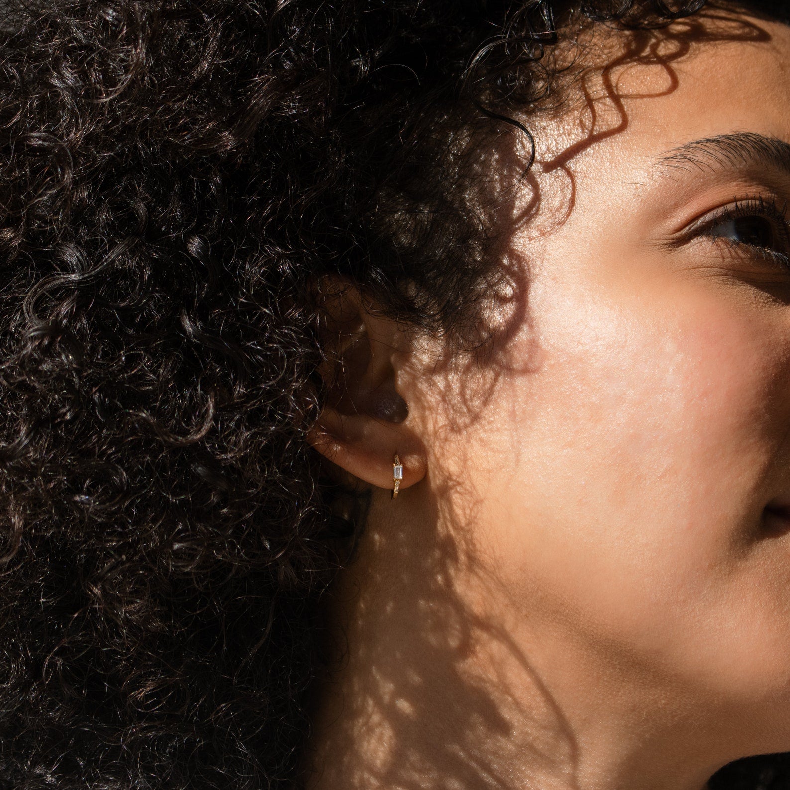 Close-up of a woman with curly hair wearing Natalie Pave Huggies, sunlight casting gentle shadows on her face.