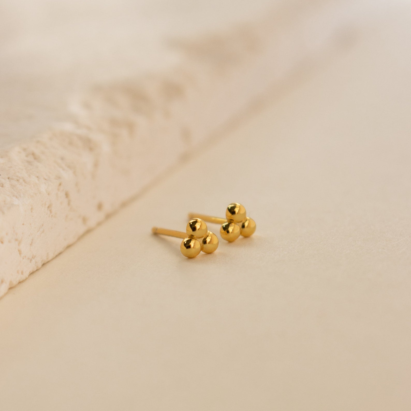 The Trio Sphere Studs are small gold earrings featuring three round beads each, ideal for earring stacks or alternative piercings, displayed on a light textured surface.