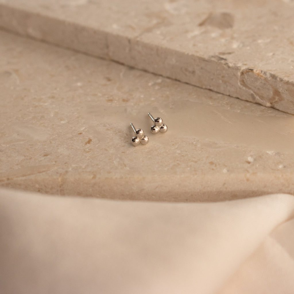 Trio Sphere Studs in silver are displayed on a beige stone surface with soft fabric in front—ideal for stacking or unique piercings.