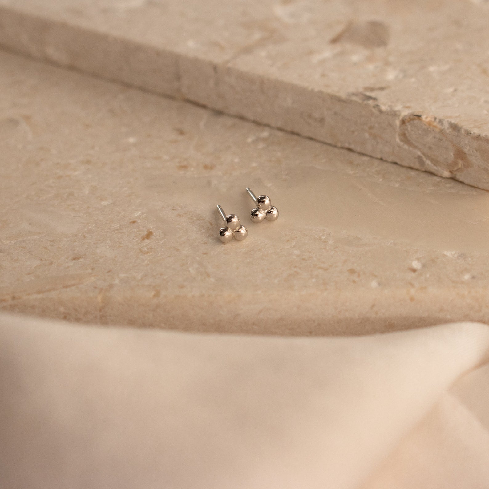 Trio Sphere Studs in silver are displayed on a beige stone surface with soft fabric in front—ideal for stacking or unique piercings.