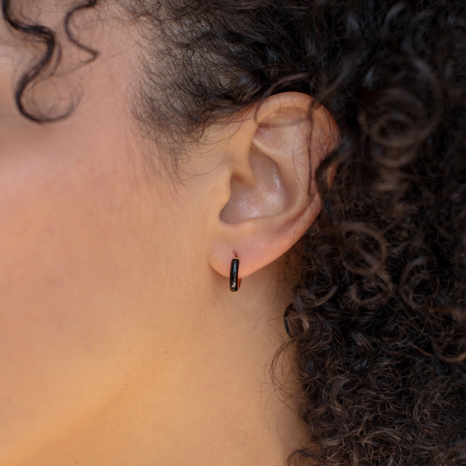 A close-up of a person with curly hair wearing Black Enamel Huggies hoop earrings.