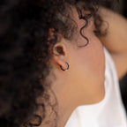 Close-up of a person with curly hair wearing the Black Enamel Huggies, small silver hoop earrings, in their ear.