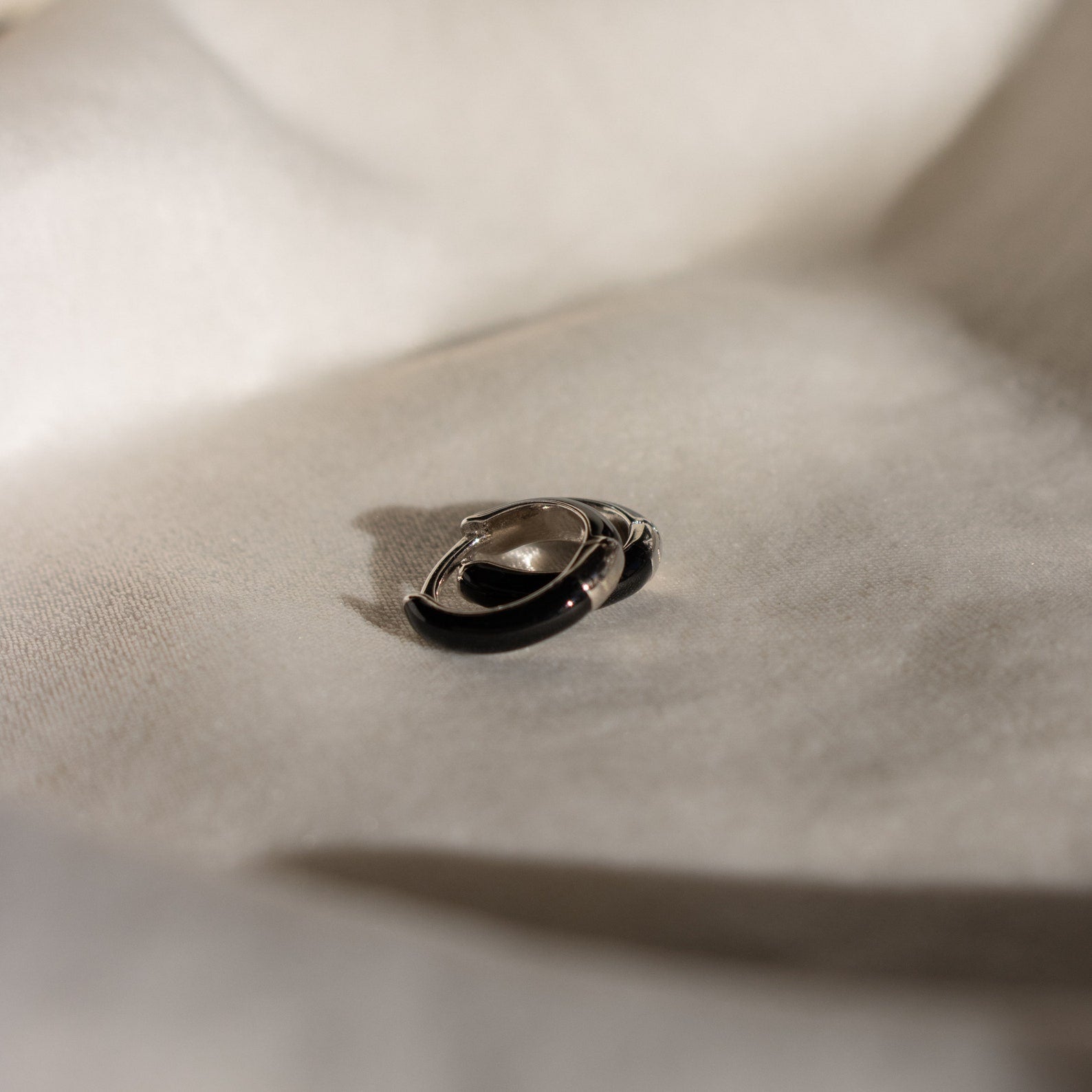 Black Enamel Huggies rest on a soft, light-colored fabric, highlighted by gentle shadows.