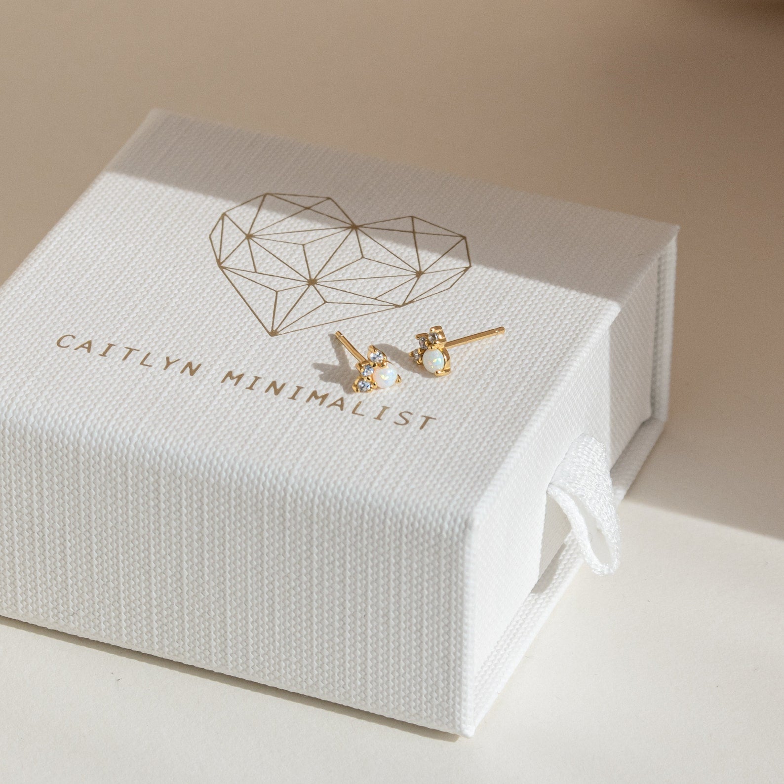 The Dainty Opal Earrings Set in gold sits on a white Caitlyn Minimalist jewelry box with a heart design, making this elegant earring set a charming gift.