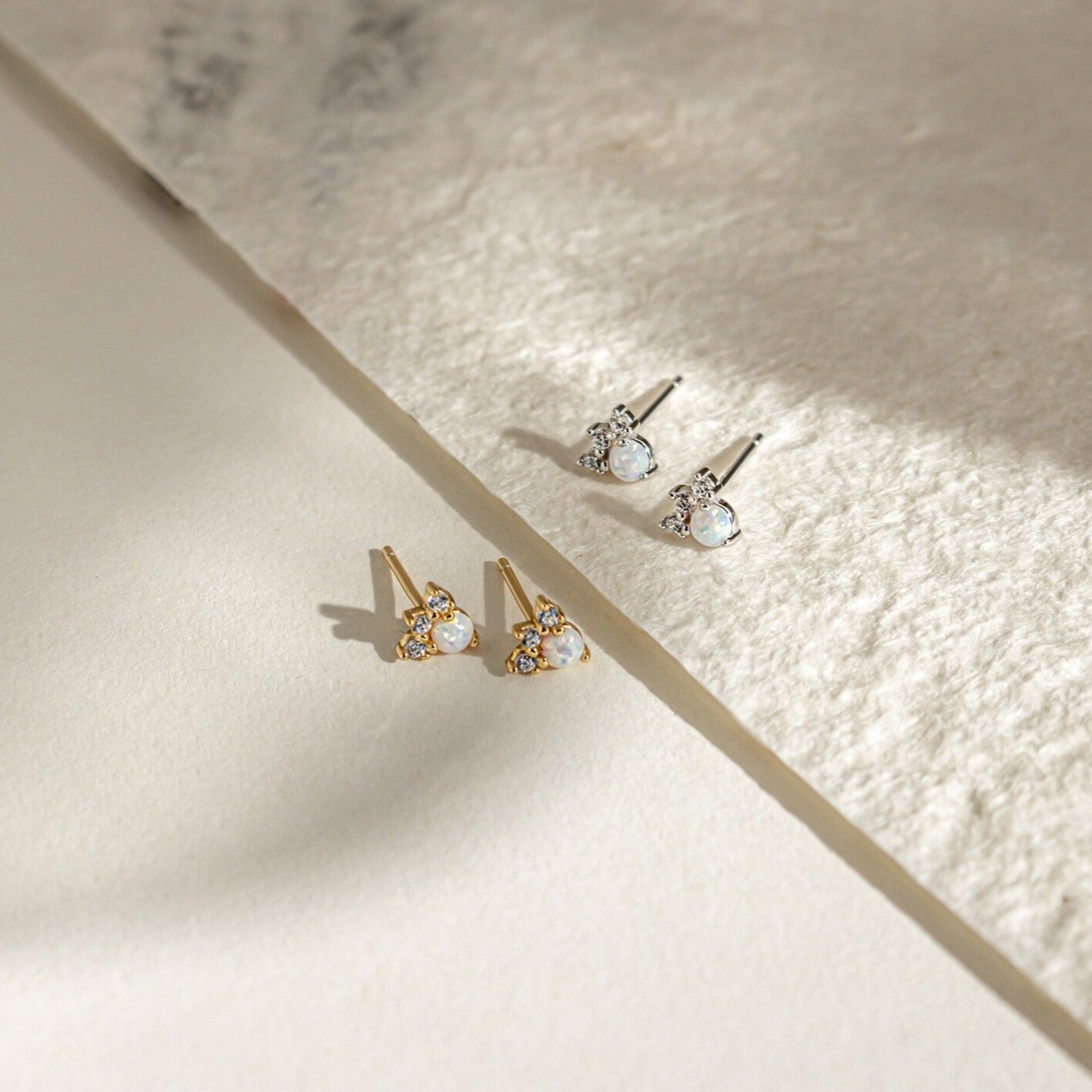 A pair of Jackie Opal Studs in gold and another in silver, each featuring small round gemstones, rest elegantly on a textured surface.