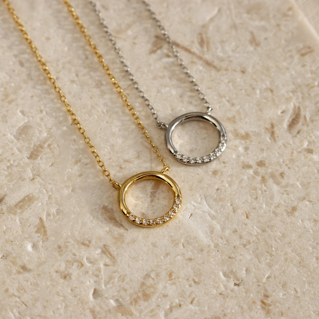 The Kayla Pave Circle Necklace, featuring gold and silver finishes with small crystals, is perfect for minimalistic jewelry lovers and is displayed on a textured beige surface.