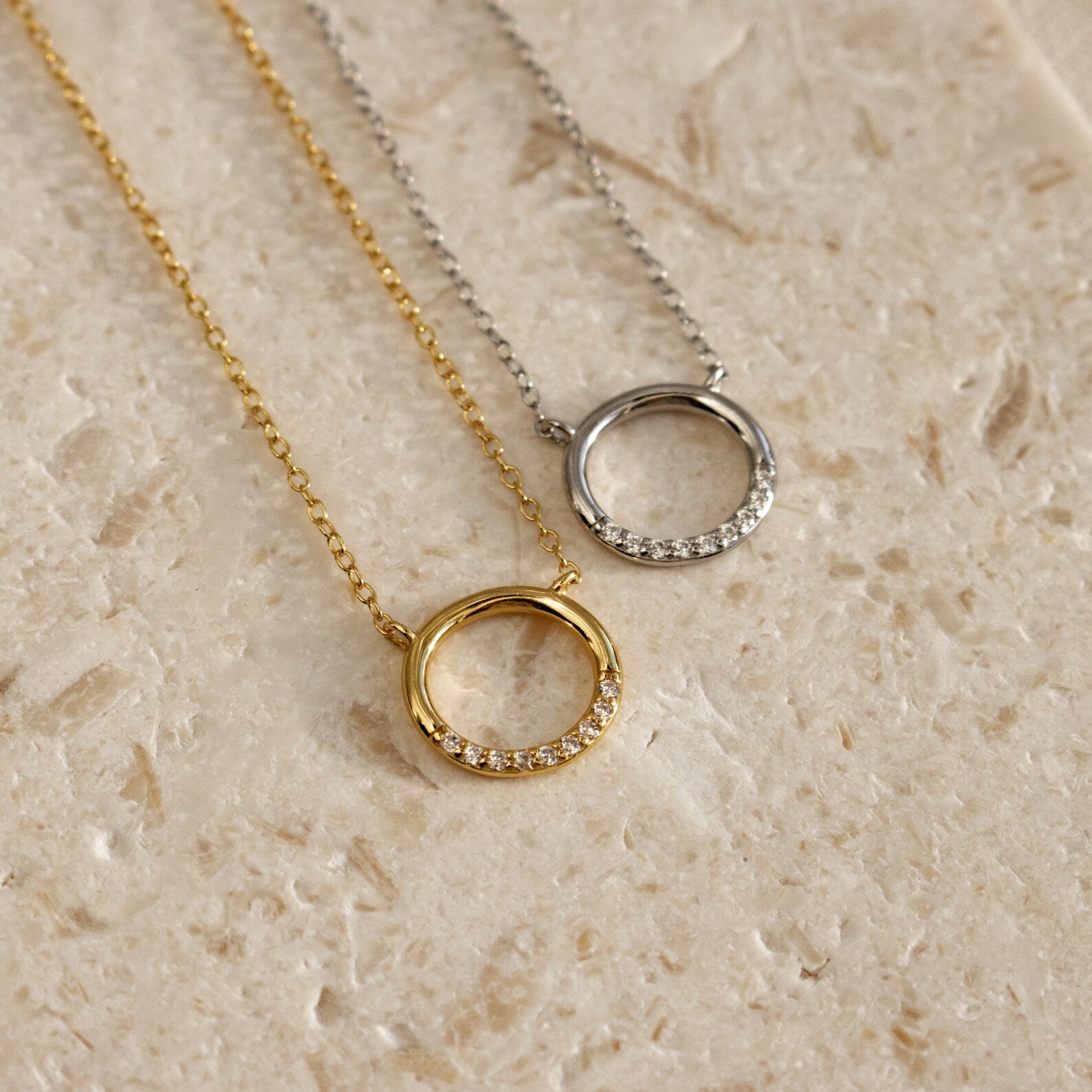 The Kayla Pave Circle Necklace, featuring gold and silver finishes with small crystals, is perfect for minimalistic jewelry lovers and is displayed on a textured beige surface.