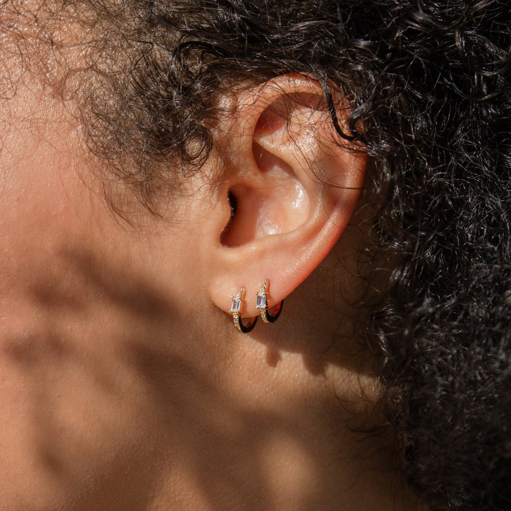Close-up of a person's ear wearing Natalie Pave Huggies—two small gold hoops, each set with a rectangular clear stone that evokes the look of baguette diamond earrings.