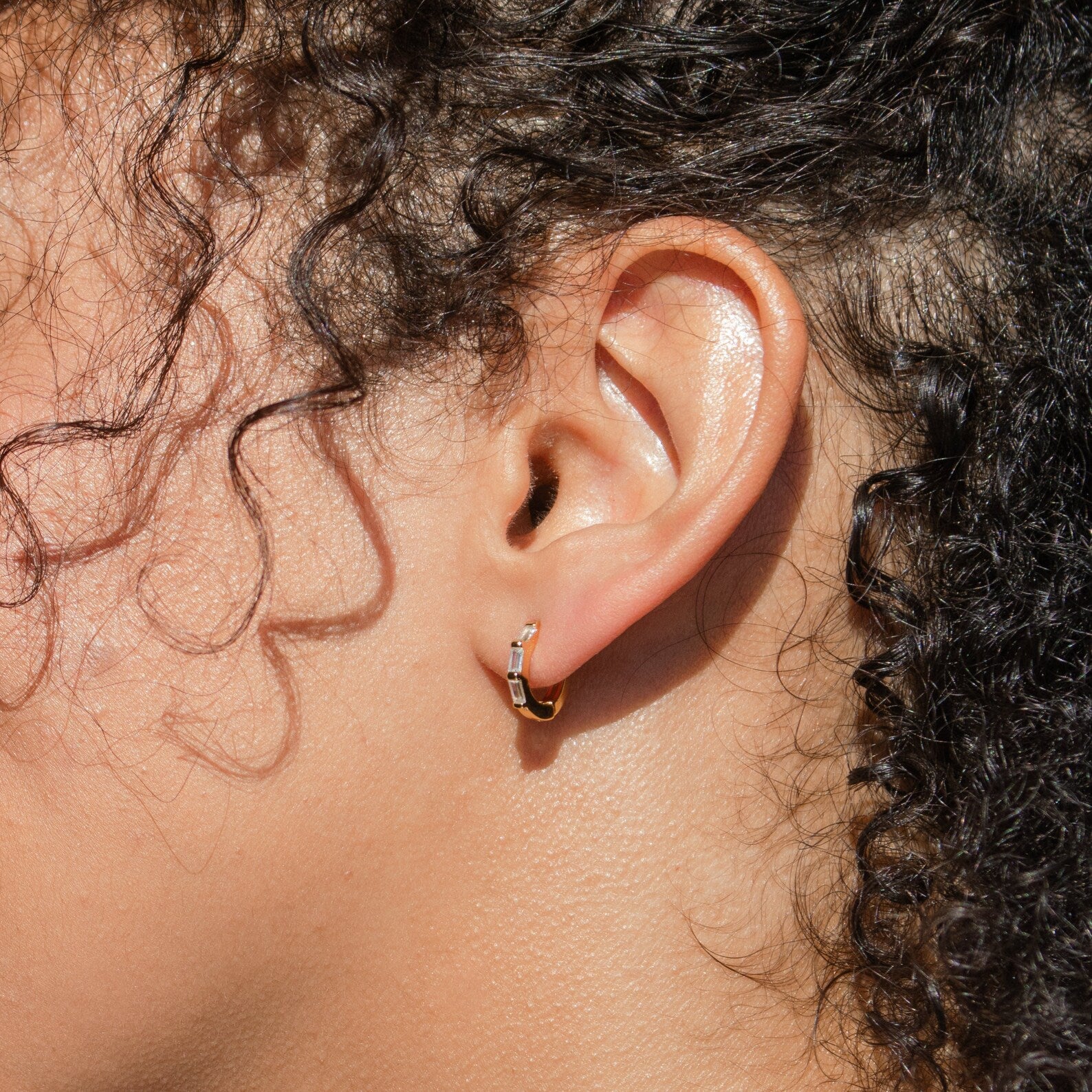 A close-up of a person's ear wearing the elegant Kenia Diamond Huggies—a small gold hoop earring—framed by curly hair.
