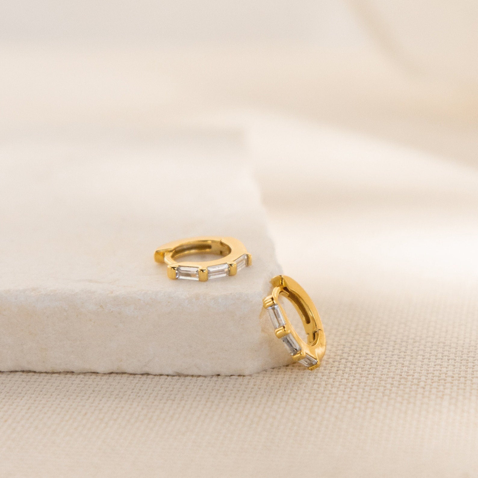 Kenia Diamond Huggies, gold hoop earrings with rectangular clear stones inspired by classic baguette diamonds, rest on a light stone surface and fabric background.