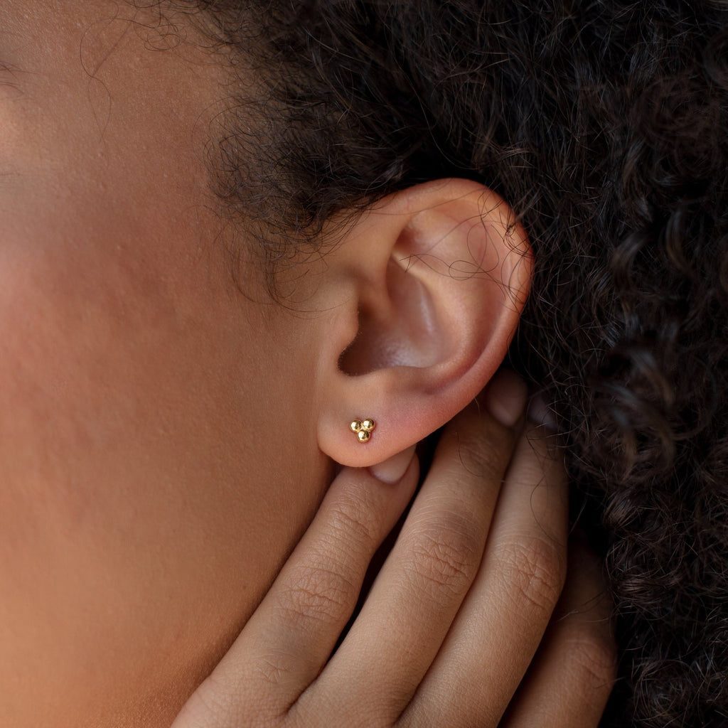 A close-up of a person’s ear showcases the Trio Sphere Studs—small gold earrings styled as part of an elegant earring stack.