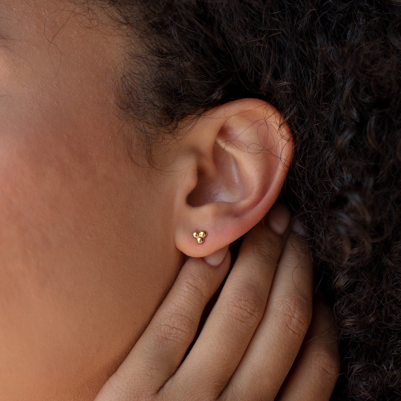 A close-up of a person’s ear showcases the Trio Sphere Studs—small gold earrings styled as part of an elegant earring stack.