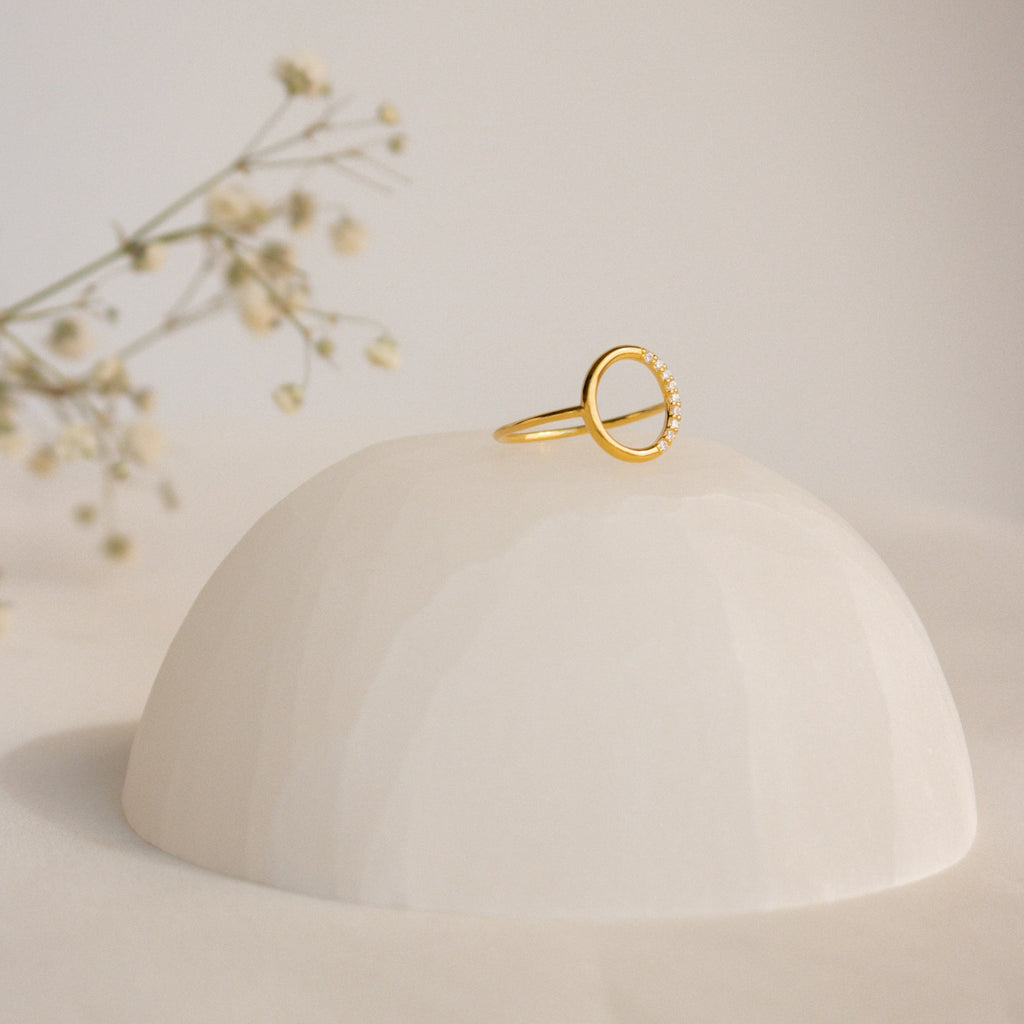The Pave Open Circle Ring features a gold band set with small gems, showcased on a smooth white dome with flower sprigs in the background for an elegant touch.
