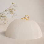 The Pave Open Circle Ring features a gold band set with small gems, showcased on a smooth white dome with flower sprigs in the background for an elegant touch.