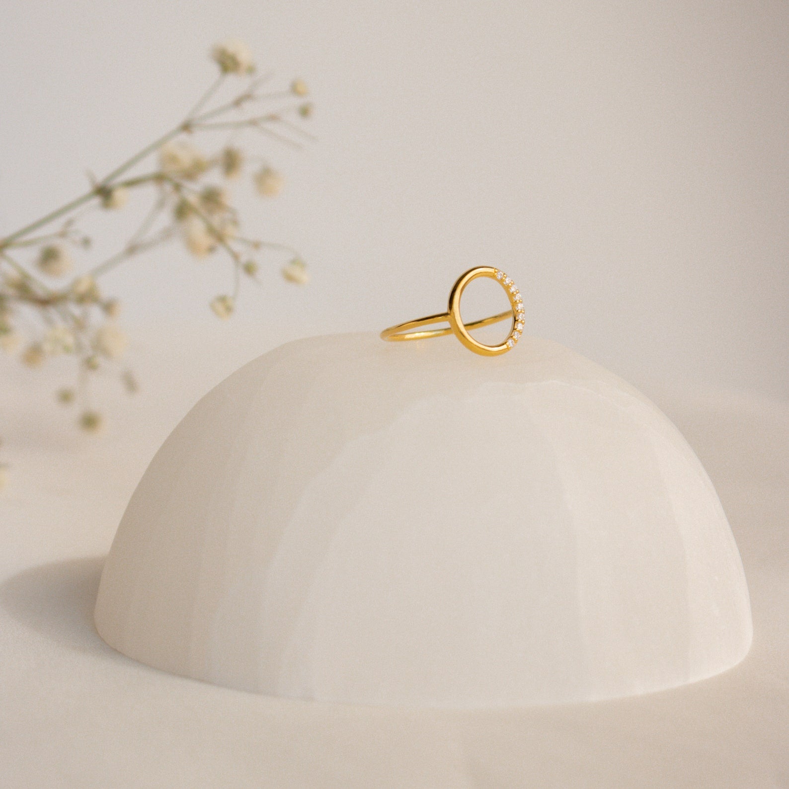 The Pave Open Circle Ring features a gold band set with small gems, showcased on a smooth white dome with flower sprigs in the background for an elegant touch.