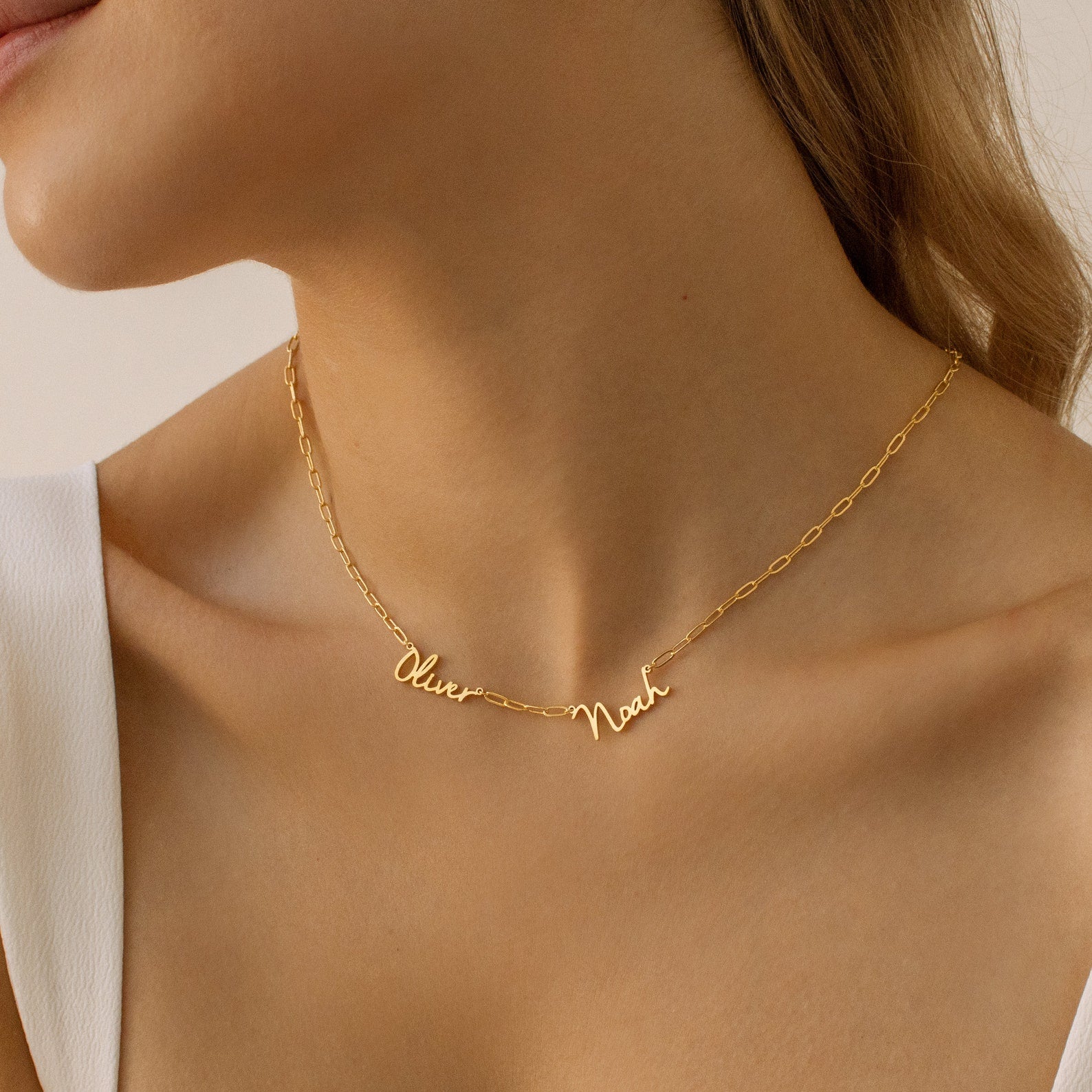 A woman wears the Nella Multiple Name Necklace in Paperclip Chain, featuring the names Oliver and Noah in cursive on a delicate gold chain.
