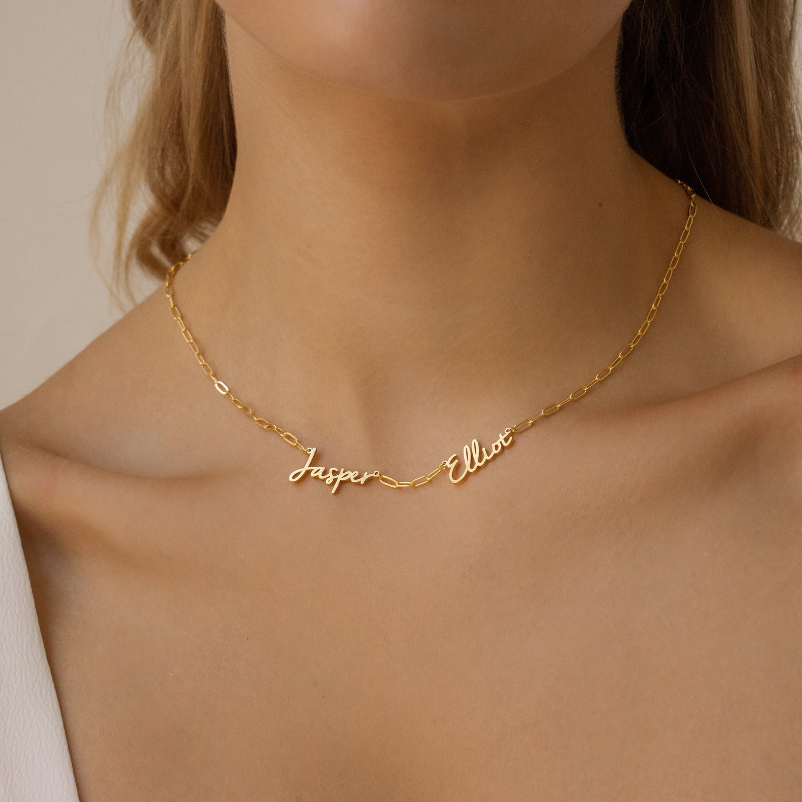 A woman wears the Nella Multiple Name Necklace in a bold paperclip chain, showcasing the names Jasper and Elliot in elegant cursive script.