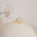 The Pave Open Circle Ring features a delicate gold band accented with small diamonds, beautifully presented on a white surface with softly blurred flowers in the background.