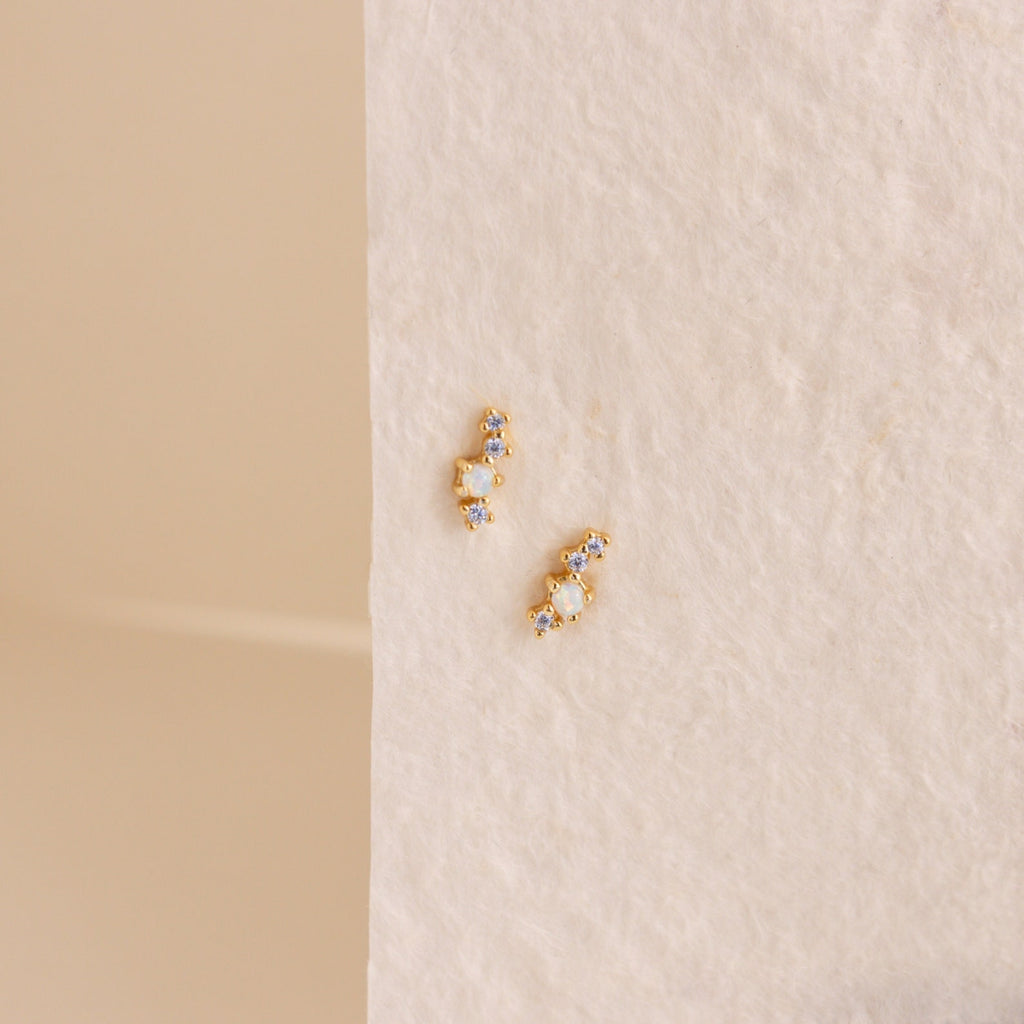 The Nile Opal and Diamond Studs, two small gold constellation-inspired earrings with sparkling stones, rest on a textured light cream surface.