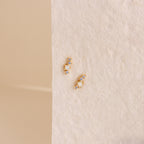 The Nile Opal and Diamond Studs, two small gold constellation-inspired earrings with sparkling stones, rest on a textured light cream surface.