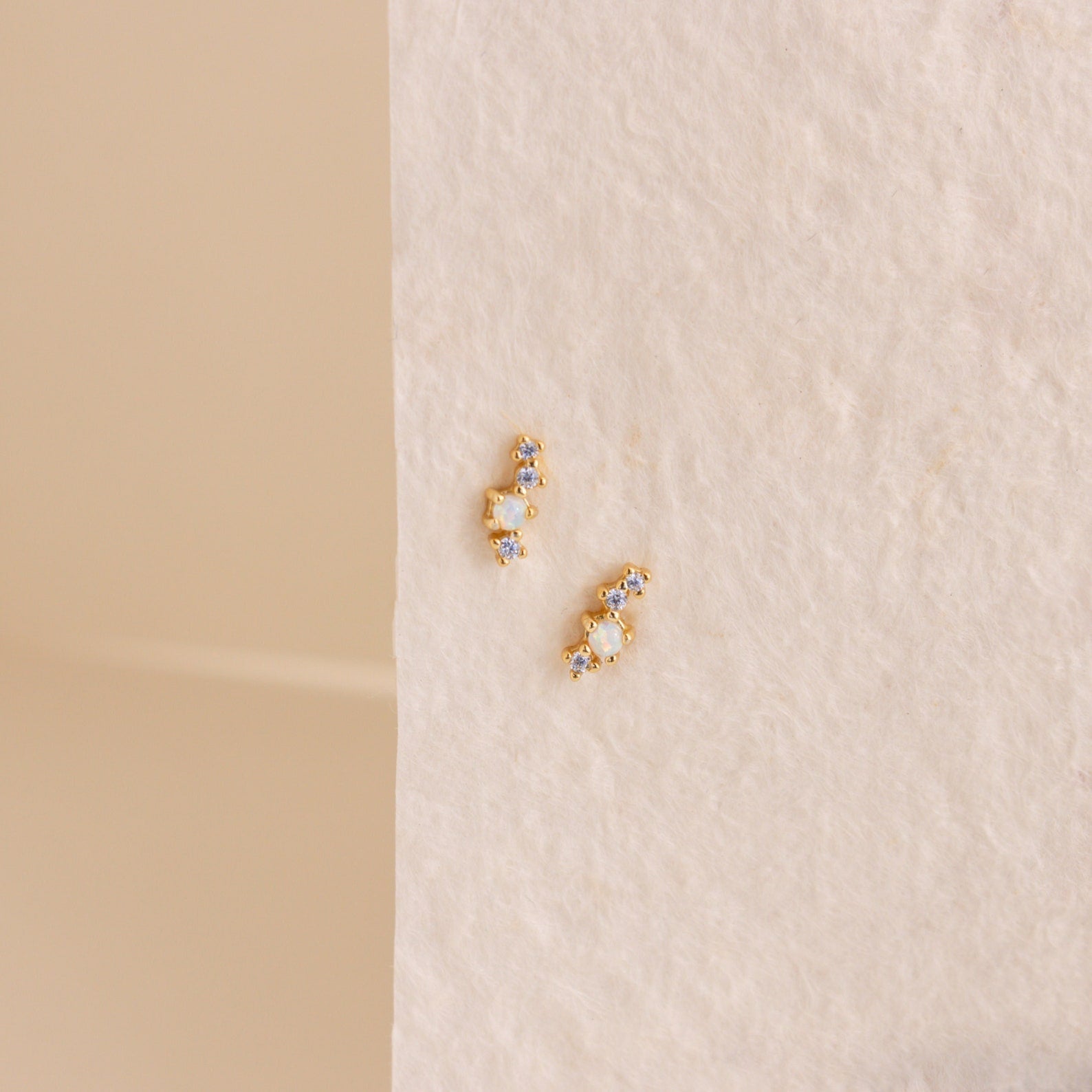 The Nile Opal and Diamond Studs, two small gold constellation-inspired earrings with sparkling stones, rest on a textured light cream surface.