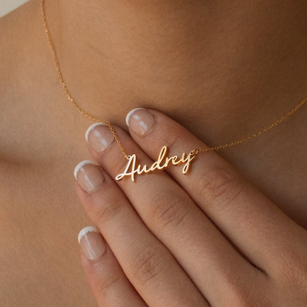 A hand with a French manicure holds the Nella Name Necklace with a gold cursive nameplate, shown on a person's neck.