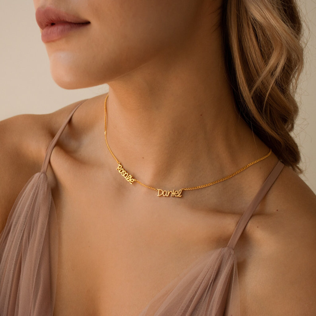 A woman wears the Preppy Multiple Name Necklace in Curb Chain with “Rebecca” and “Daniel” in a youthful font on gold, paired with a sheer taupe dress.