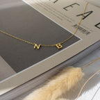The Alice Dainty Initial Necklace, with letter charms N and B, rests on a beige magazine beside dried wheat—a personalized jewelry piece perfect for Christmas gifting.