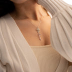 A woman models the Flower Name Necklace, featuring a personalized name pendant and a silver key-shaped charm, styled with a cream cardigan and tank top.