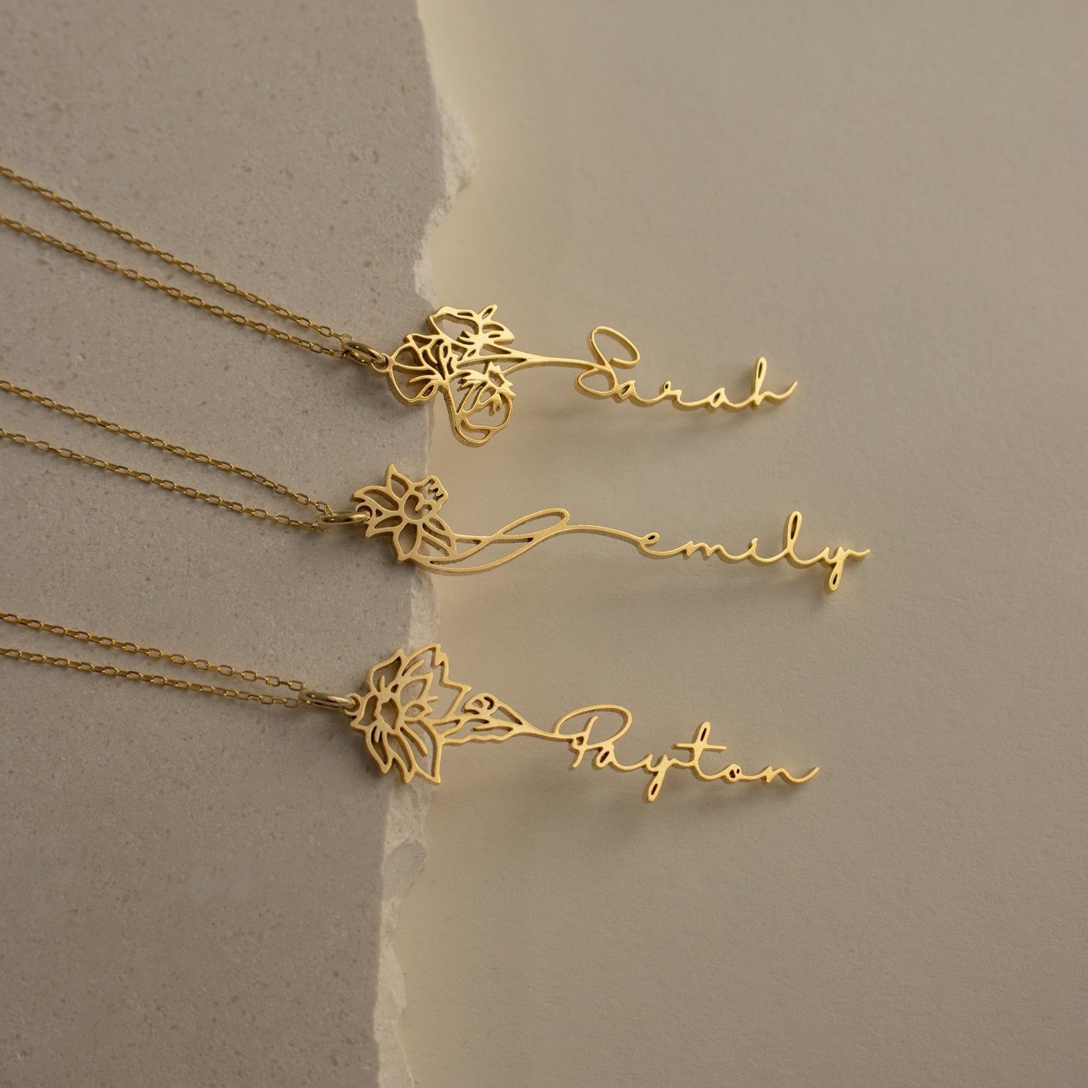Three gold Flower Name Necklaces, each with a unique floral design and personalized with the names Sarah, Emily, and Payton plus their birth flowers, are displayed on a neutral background.