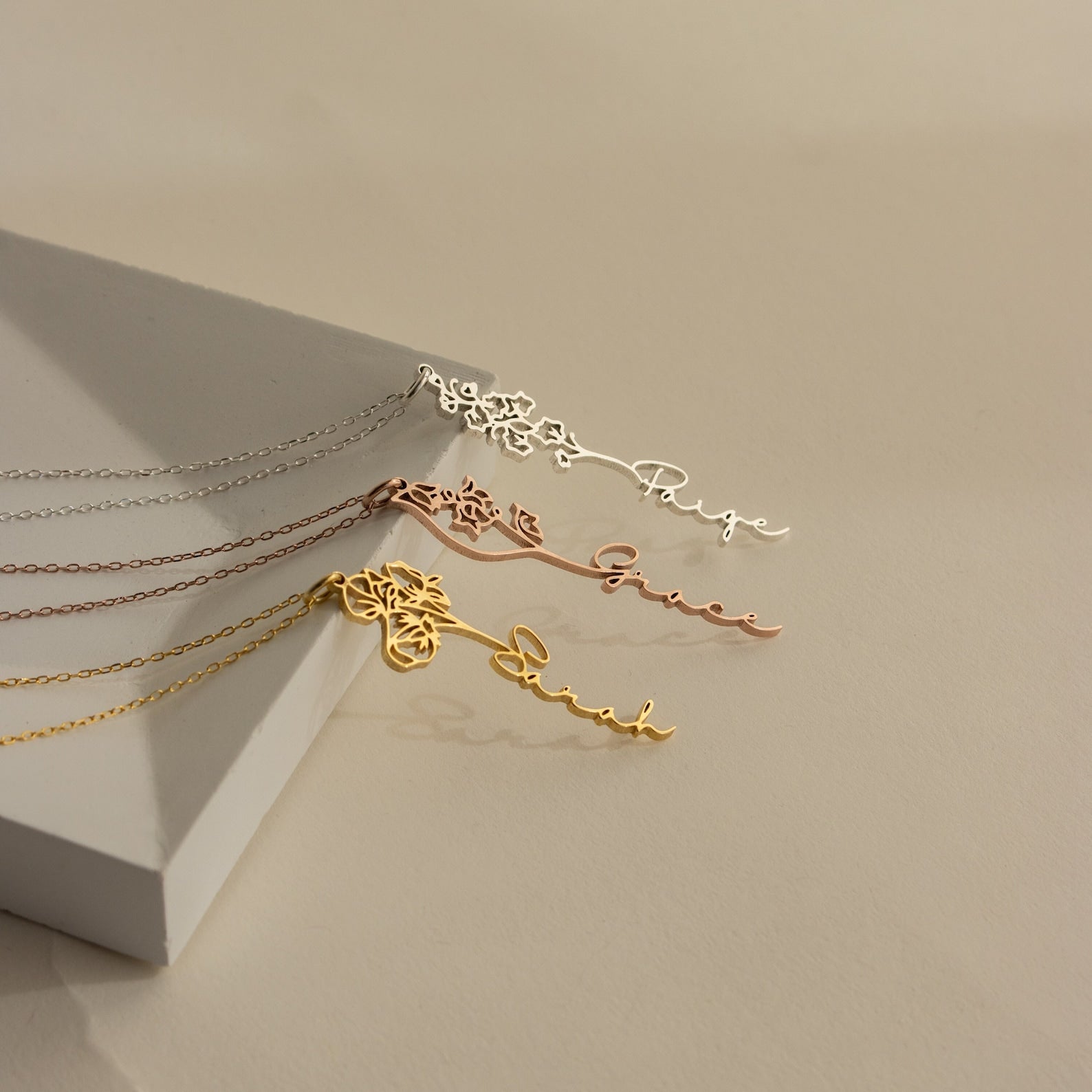 Three Flower Name Necklaces in silver, rose gold, and gold—each personalized with a name and delicate birth flowers—are displayed on a beige background.