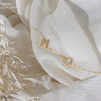 Gold bracelet with letter M and a circular charm on white textured fabric, surrounded by dried florals—a dainty piece that pairs perfectly with our Pave Sideways Initial Necklace for a personalized look.