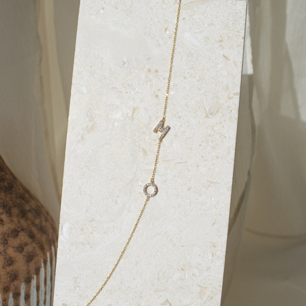 The Pave Sideways Initial Necklace showcases a diamond letter "N" and a circular pendant, elegantly displayed on a light textured stone background.