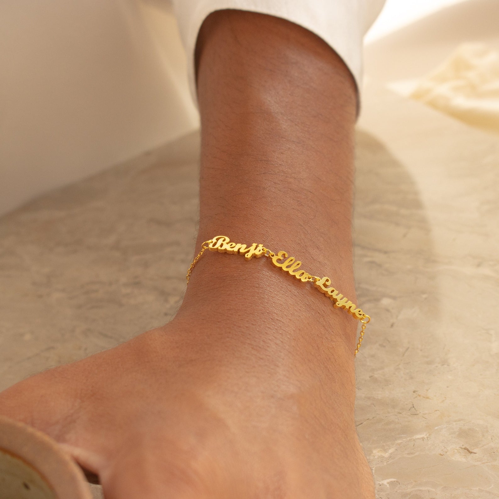 The London Multiple Name Bracelet, featuring gold cursive lettering, is displayed elegantly on a marble surface—adding a touch of personalized dainty jewelry charm to any wrist.