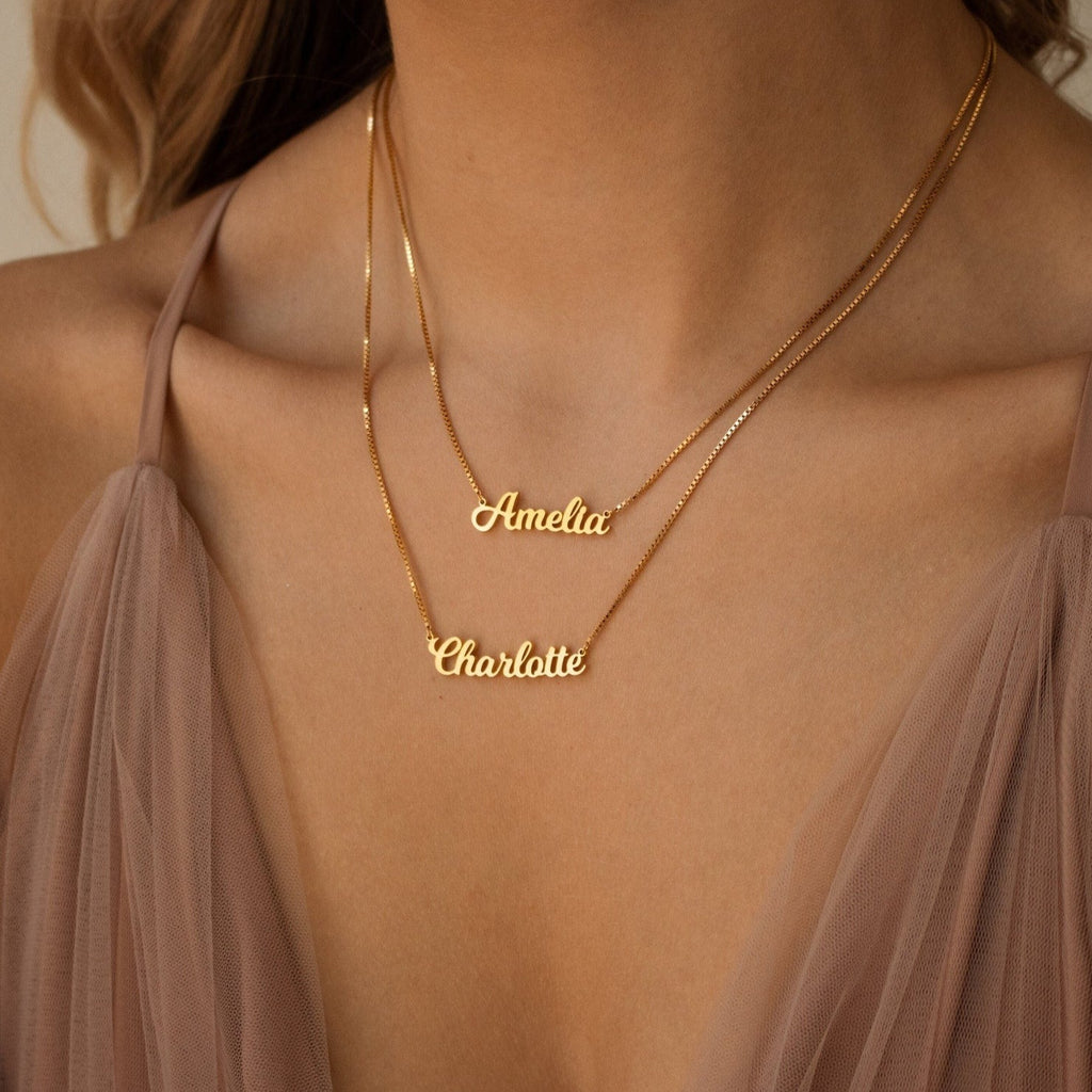 Wearing a mauve dress, a woman showcases the Mellow Double Name Necklace in Box Chain, personalized with “Amelia” and “Charlotte” in a classic font.