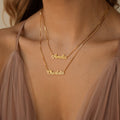 Wearing a mauve dress, a woman showcases the Mellow Double Name Necklace in Box Chain, personalized with “Amelia” and “Charlotte” in a classic font.