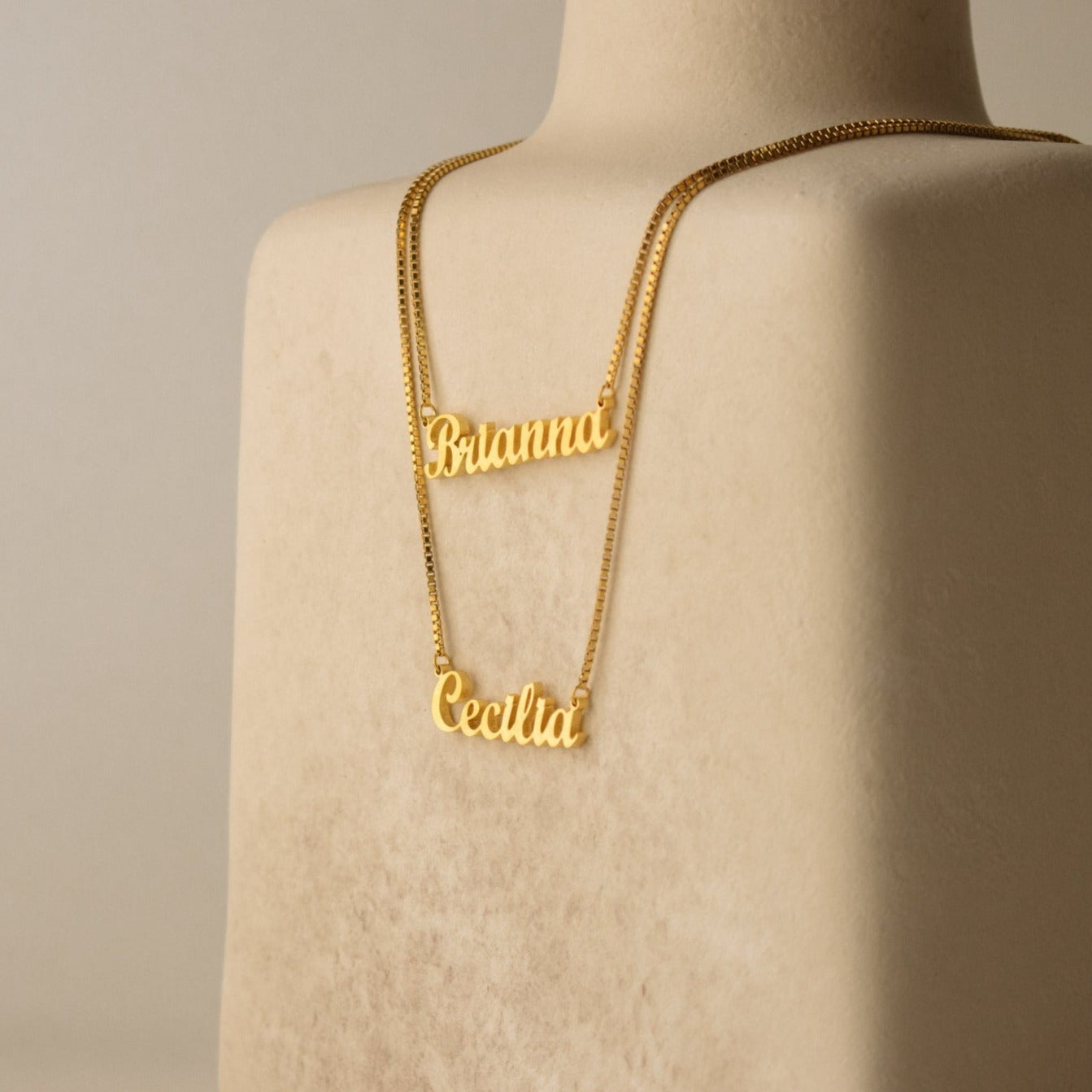 The London Double Name Necklace in Box Chain features two chic name necklaces, displayed on a beige bust and highlighting the charm of personalized jewelry in an elegant double-name design.