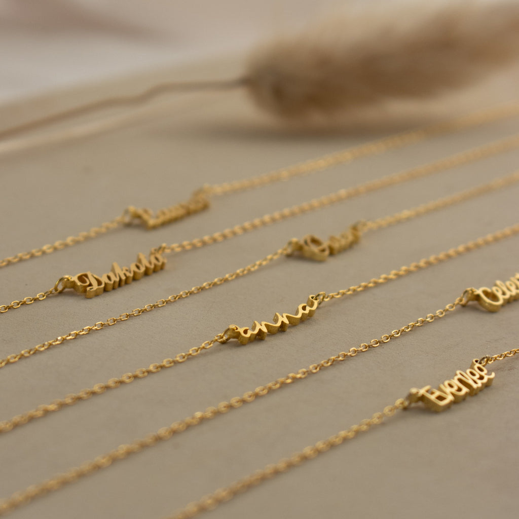 The Tiny Summer Name Necklace features a delicate gold chain with a custom name pendant in an elegant font, displayed on a beige surface.