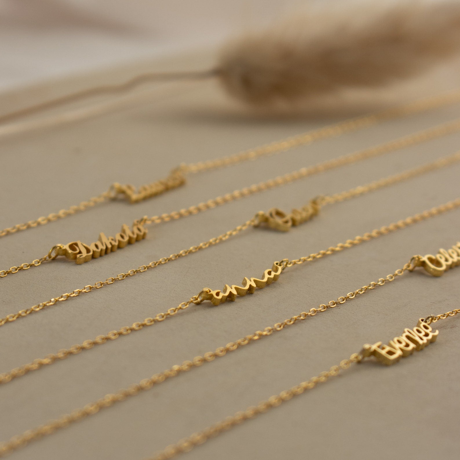 The Tiny Summer Name Necklace features a delicate gold chain with a custom name pendant in an elegant font, displayed on a beige surface.